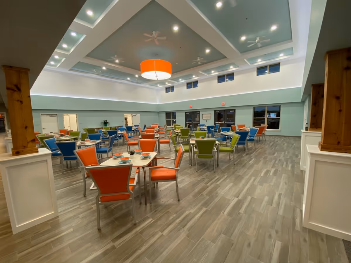 Spacious, brightly lit dining room with colorful orange, green, and blue chairs arranged around tables under a high coffered ceiling.