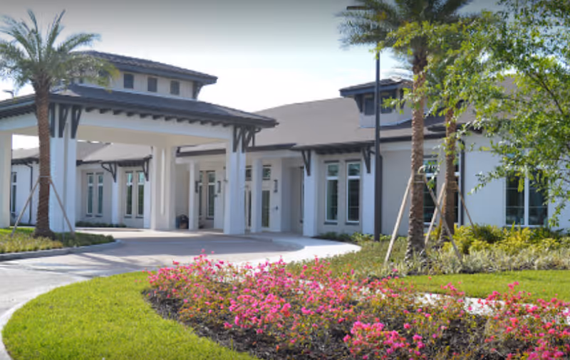 Exterior view of a senior living facility with a covered entrance, white walls, multiple windows, palm trees, and landscaped garden beds with pink flowers and green grass.