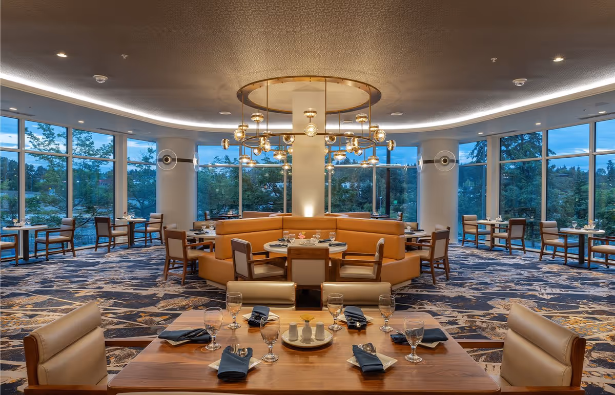 A spacious and elegant dining room with large floor-to-ceiling windows offering a view of trees outside. The room features a central circular booth with tan leather seating and multiple round tables with chairs arranged around the perimeter. The ceiling has a modern circular chandelier with multiple light fixtures. Tables are set with glassware, napkins, and silverware.