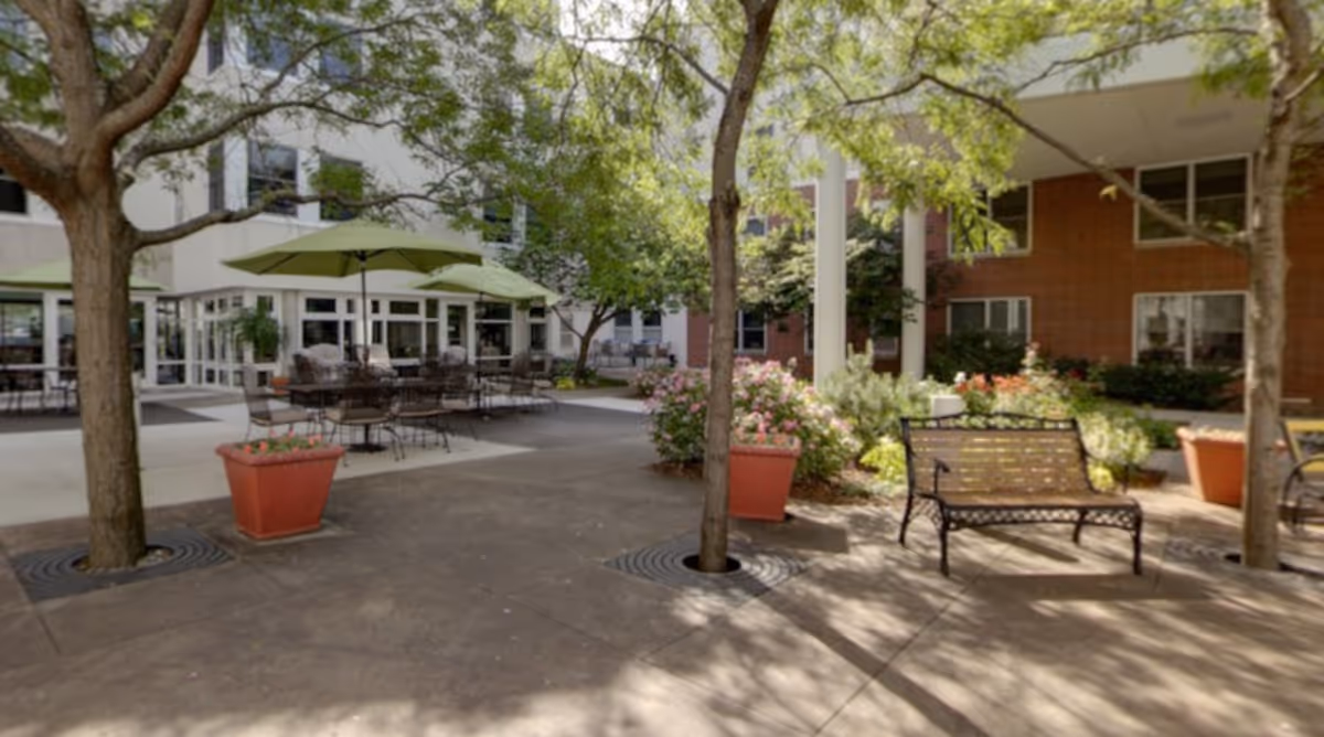 Shaded courtyard with tables and umbrellas, benches, potted plants, and surrounding building façades.