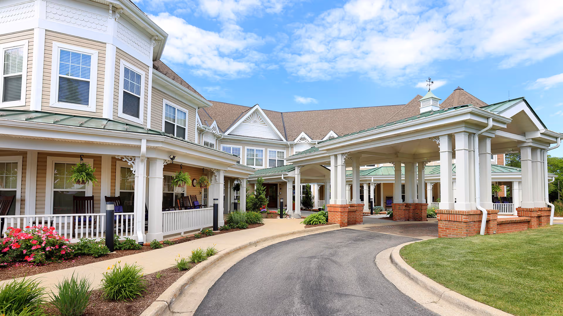 Exterior view of a senior living facility with a covered entrance driveway, white columns, brick accents, and a well-maintained garden with flowers and shrubs under a blue sky with some clouds.