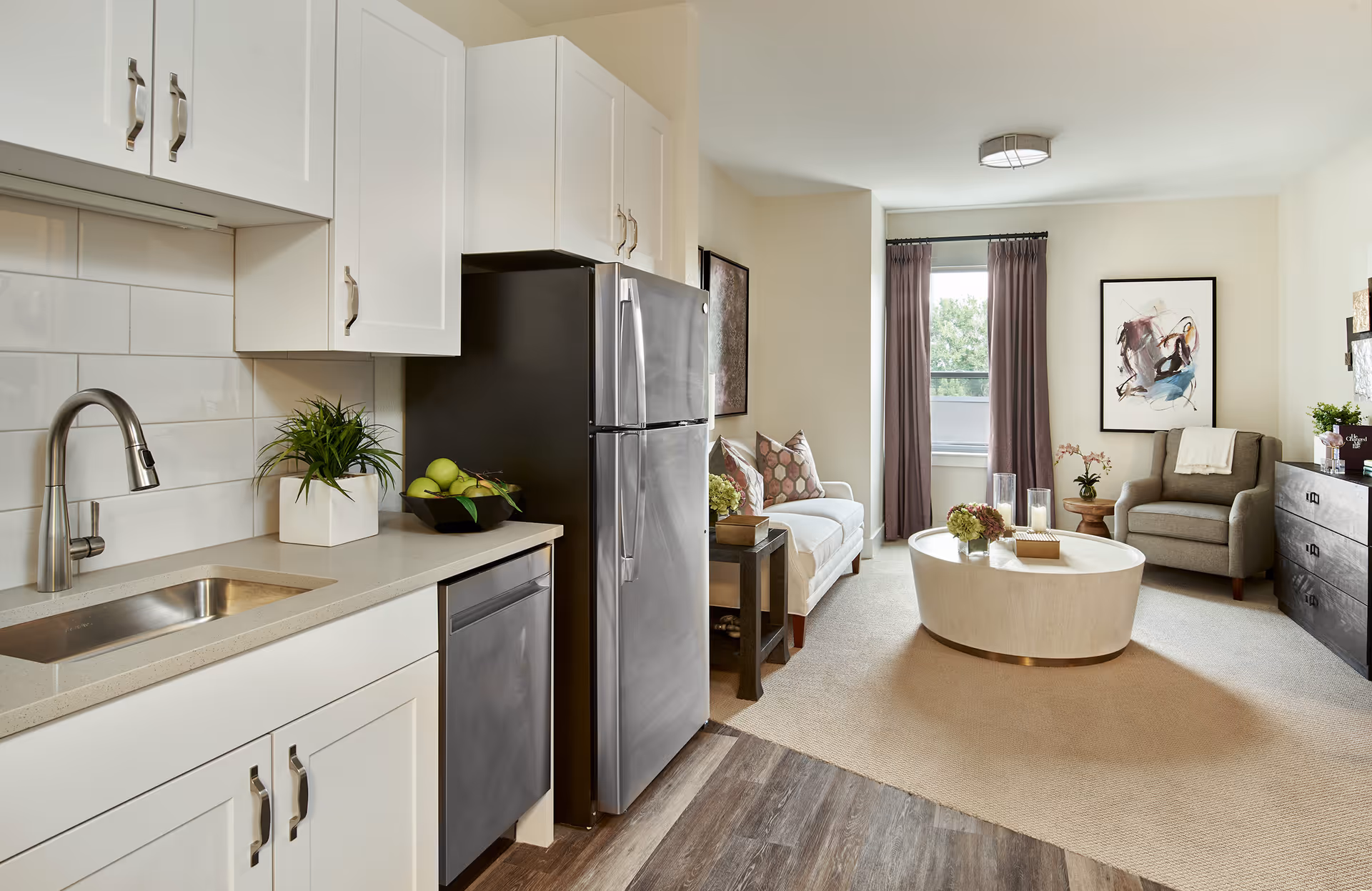 Open-plan interior showing a small kitchen with stainless steel appliances opening into a living area with a sofa, armchair, round coffee table and window with curtains.