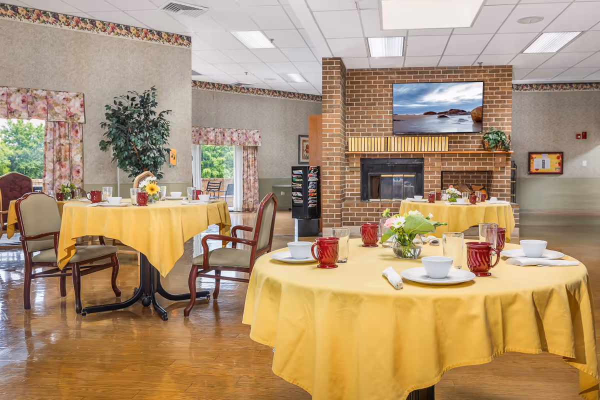 A dining room with round tables covered in yellow tablecloths, set with white plates, bowls, red glasses, and napkins. The room features wooden chairs, a brick fireplace with a flat-screen TV mounted above it, floral curtains on the windows, and a potted plant near the wall.