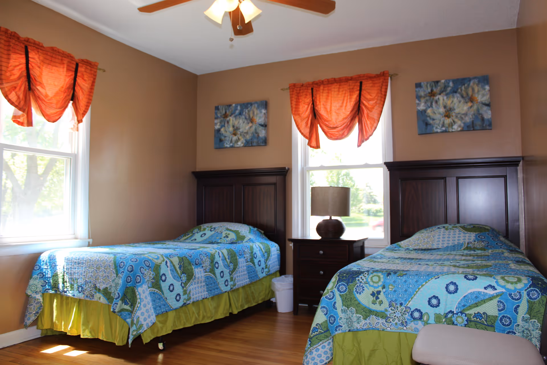 A bedroom with two twin beds featuring blue and green patterned bedspreads. Between the beds is a dark wooden nightstand with a lamp and a small trash bin underneath. The walls are painted brown and decorated with two floral paintings above each bed. Two windows with orange curtains allow natural light into the room. A ceiling fan with wooden blades is visible on the ceiling.