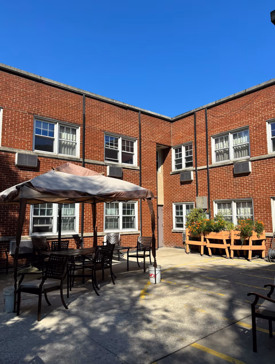 Outdoor courtyard area with a canopy-covered table and several chairs on a concrete surface. The courtyard is surrounded by a two-story red brick building with multiple windows. There are planter boxes with flowers on one side and a clear blue sky overhead.