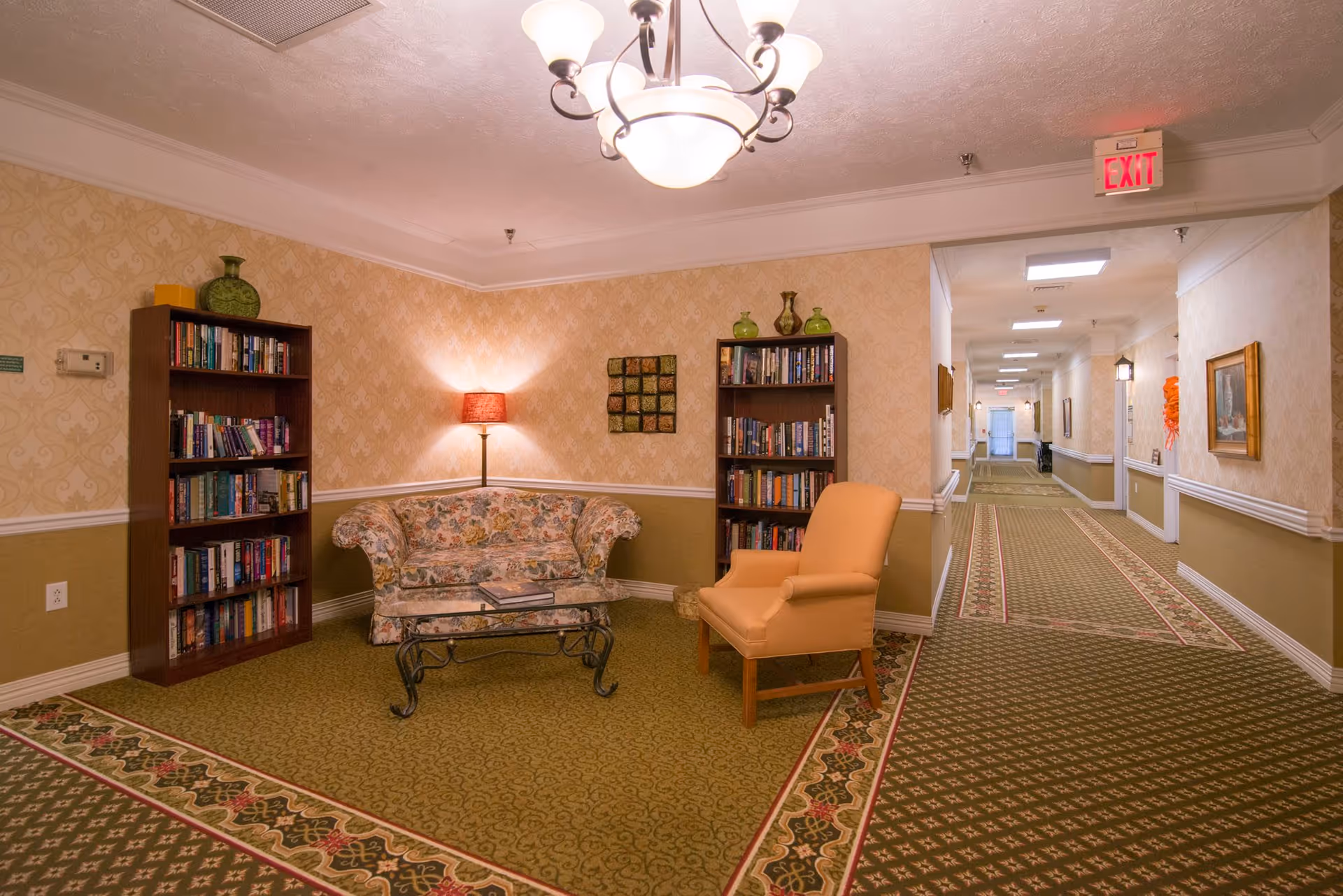 A cozy sitting area in a hallway of a senior living facility featuring a floral patterned couch, an orange armchair, two wooden bookshelves filled with books and decorative vases, a glass coffee table, a floor lamp with a red shade, and patterned carpet. The hallway extends with framed pictures on the walls and an exit sign visible at the end.