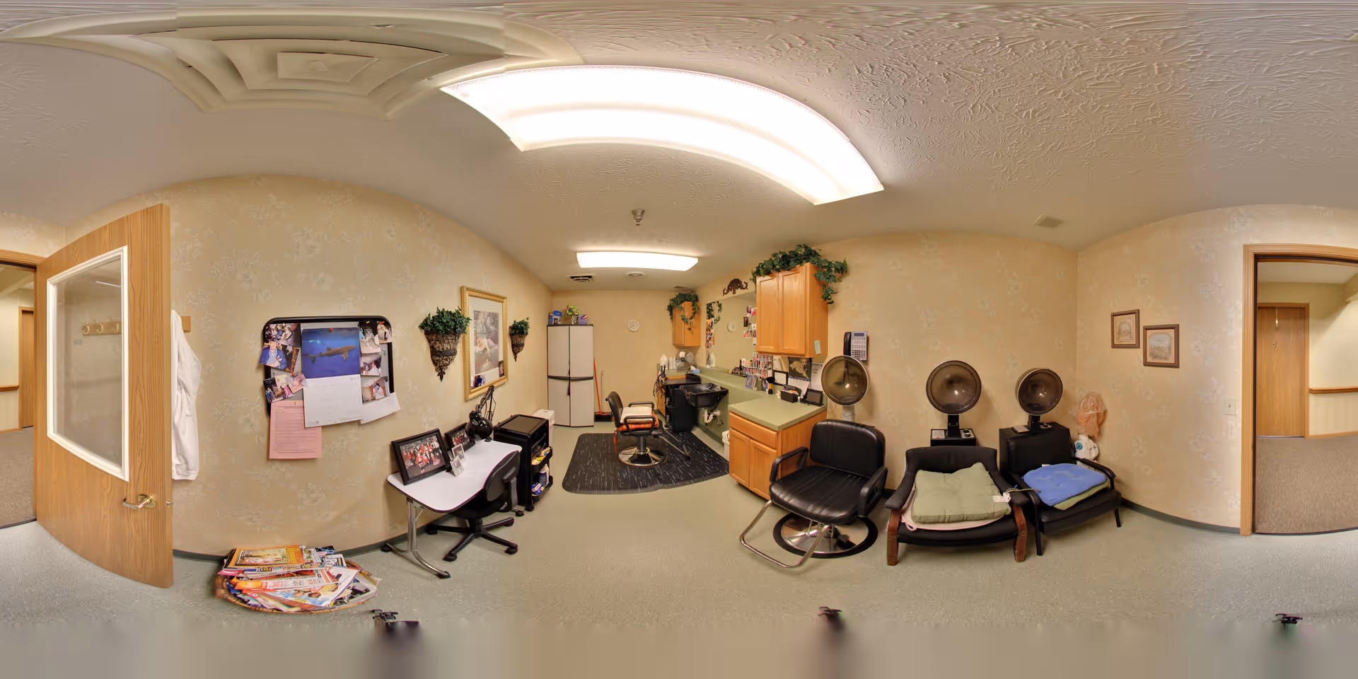 Interior view of a hair salon area within a senior living facility. The room features salon chairs, hair dryers, a workstation with a desk and chair, a refrigerator, and cabinets. The walls are decorated with framed pictures, a bulletin board with photos and notes, and some greenery above the cabinets. There are two open doors leading to adjacent rooms or hallways.