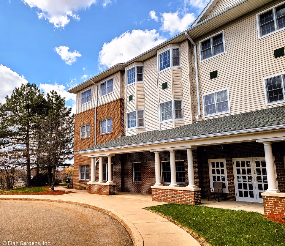 Exterior view of a multi-story senior living facility building with brick and beige siding, white columns supporting a covered entrance, and several windows. There is a curved driveway and some trees under a partly cloudy blue sky.