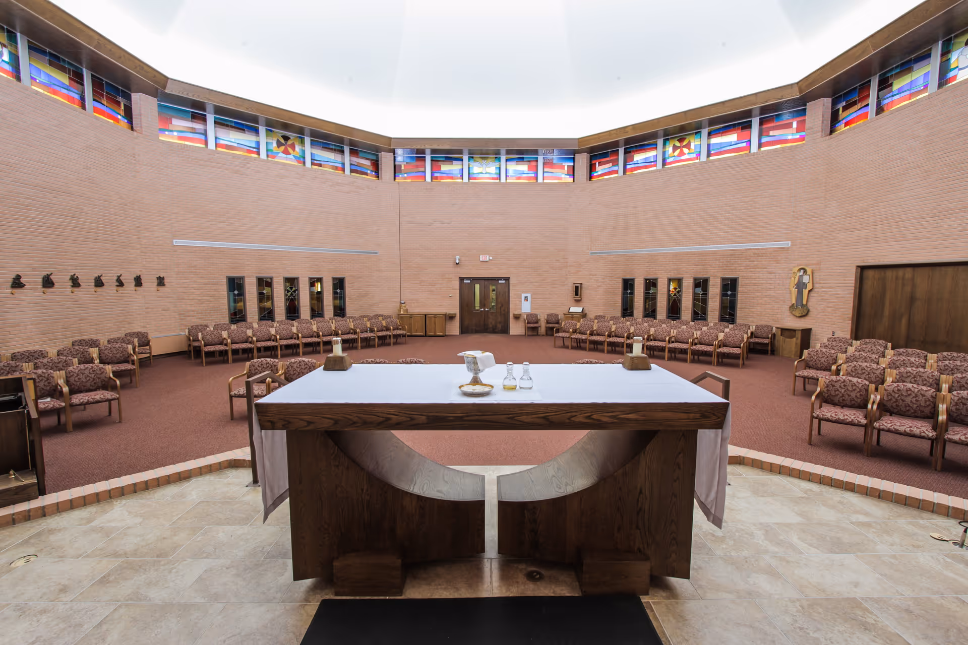 Interior of a circular chapel with an altar in the foreground, rows of chairs around the perimeter, and stained-glass clerestory windows near the ceiling.