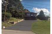 View of a residential-style building with a driveway leading up to a garage. The building is surrounded by green grass, trees, and bushes under a partly cloudy sky.