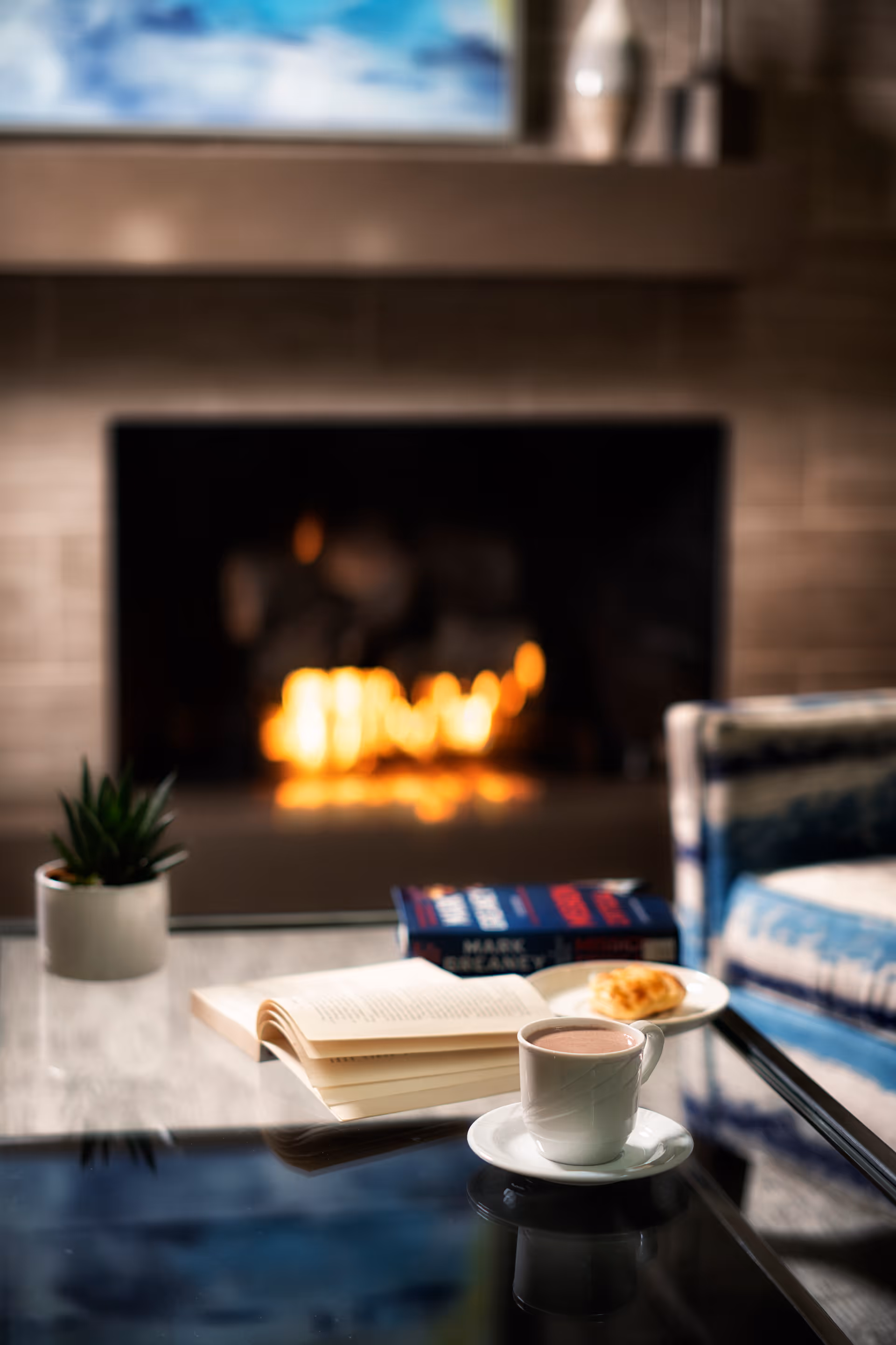 A cozy living room scene with a lit fireplace in the background, a glass coffee table in the foreground holding an open book, a cup of hot beverage on a saucer, a small plate with a pastry, a potted plant, and a closed book. A patterned armchair is partially visible to the right.