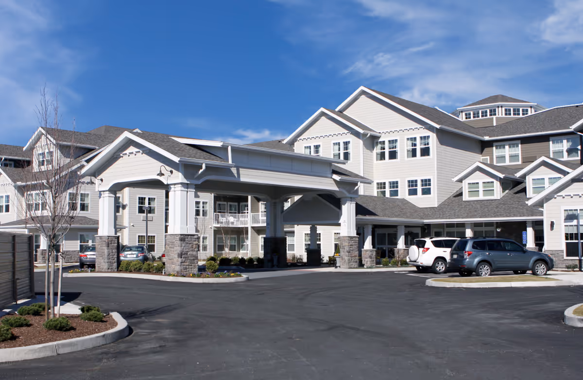 Front entrance of a multi-story senior living facility with a covered porte-cochere and parked cars under a blue sky.
