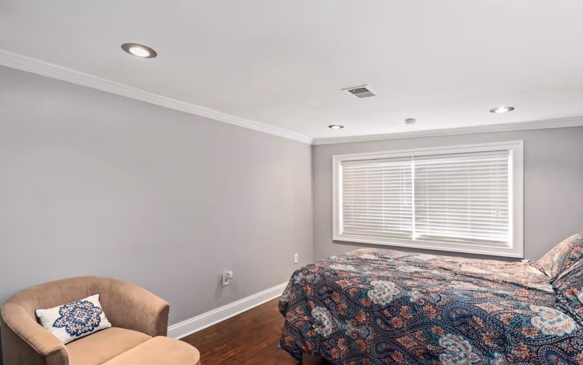 A bedroom with a bed covered in a patterned quilt, a beige armchair with a decorative pillow, hardwood floors, gray walls, a large window with white blinds, and recessed ceiling lights.