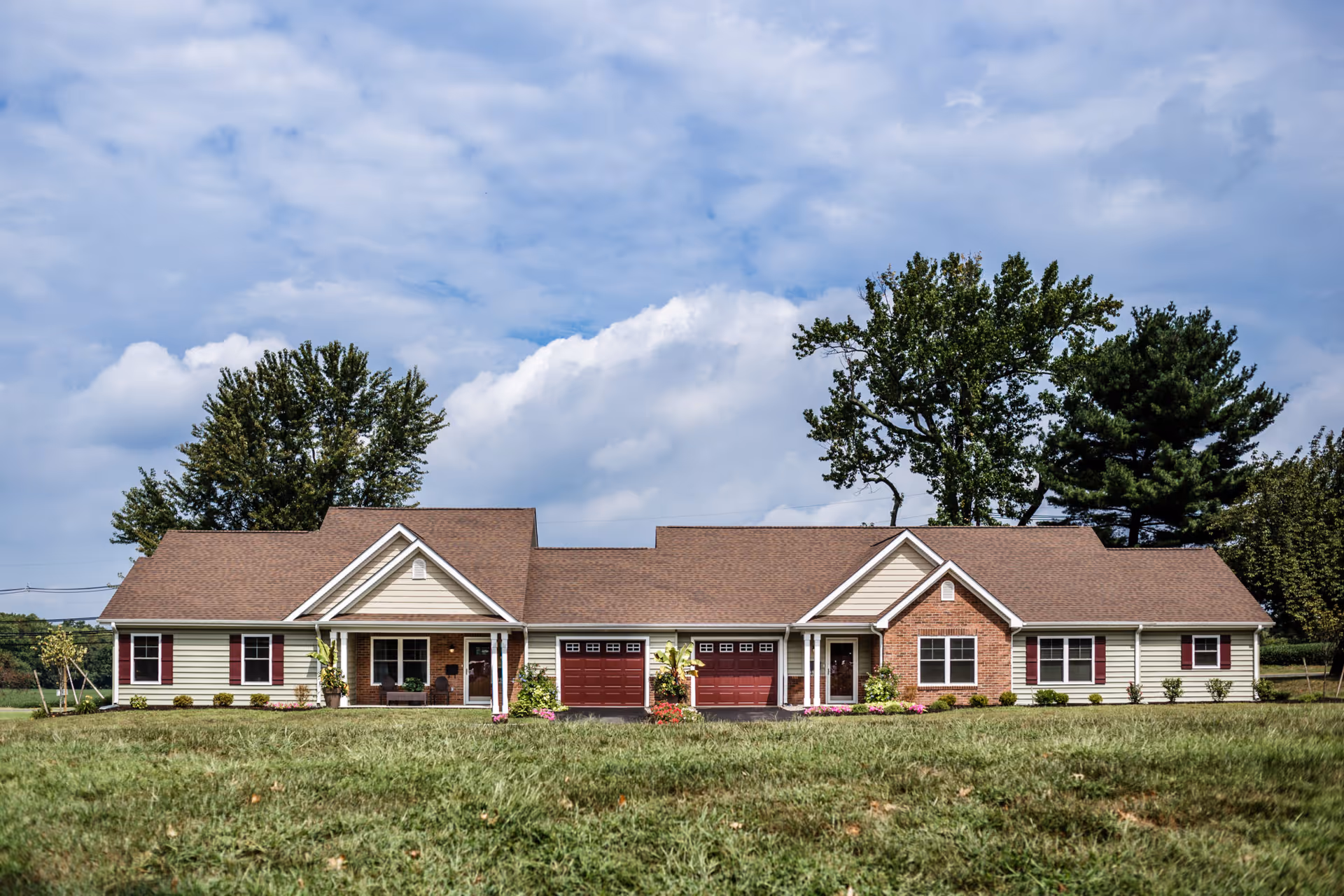 Single-story duplex-style building with two attached garages, front porches, and a large lawn under a cloudy sky.