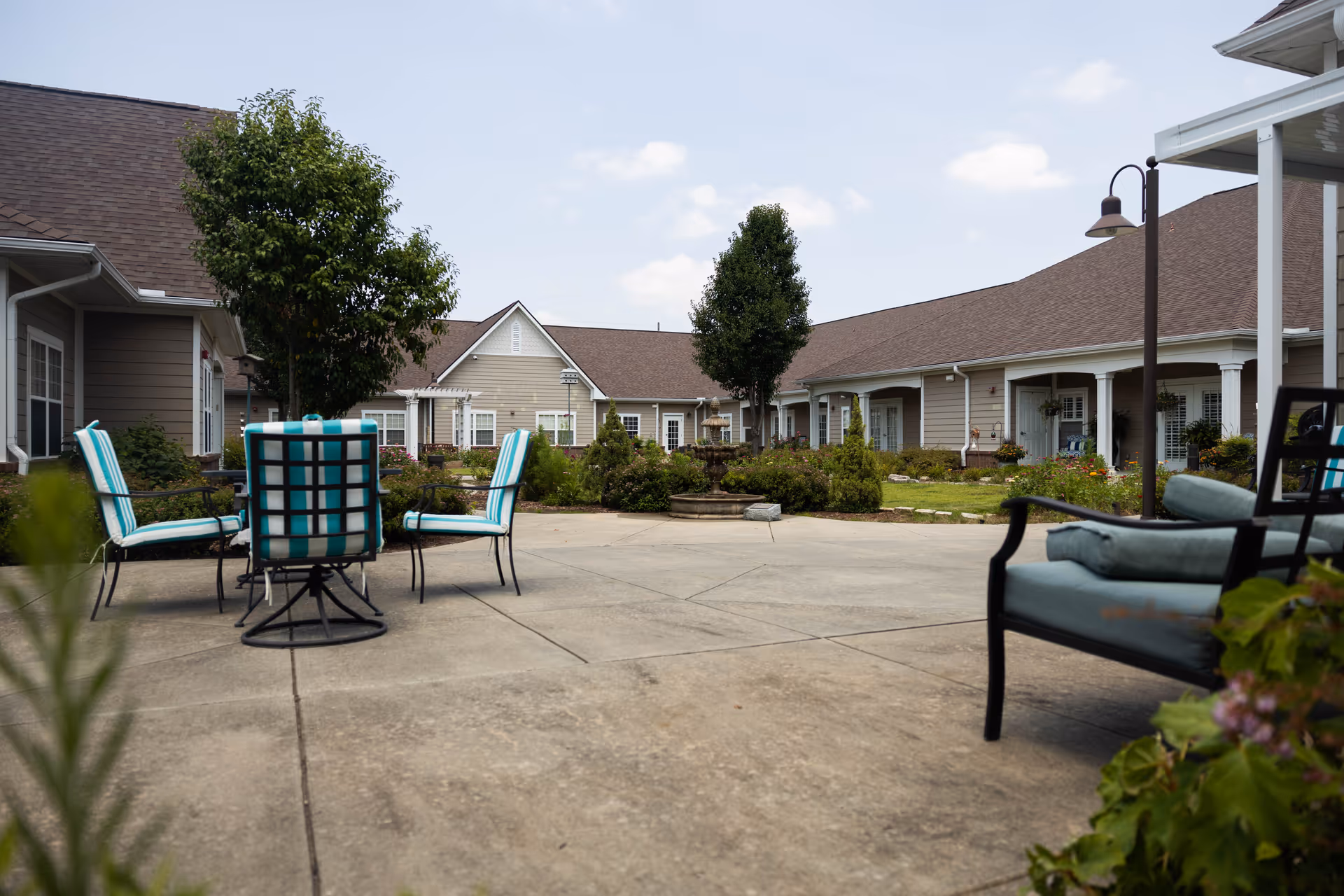 Outdoor patio area at Chateau On the Ridge with cushioned chairs around a table on a concrete surface, surrounded by greenery and trees, with beige buildings in the background under a partly cloudy sky.