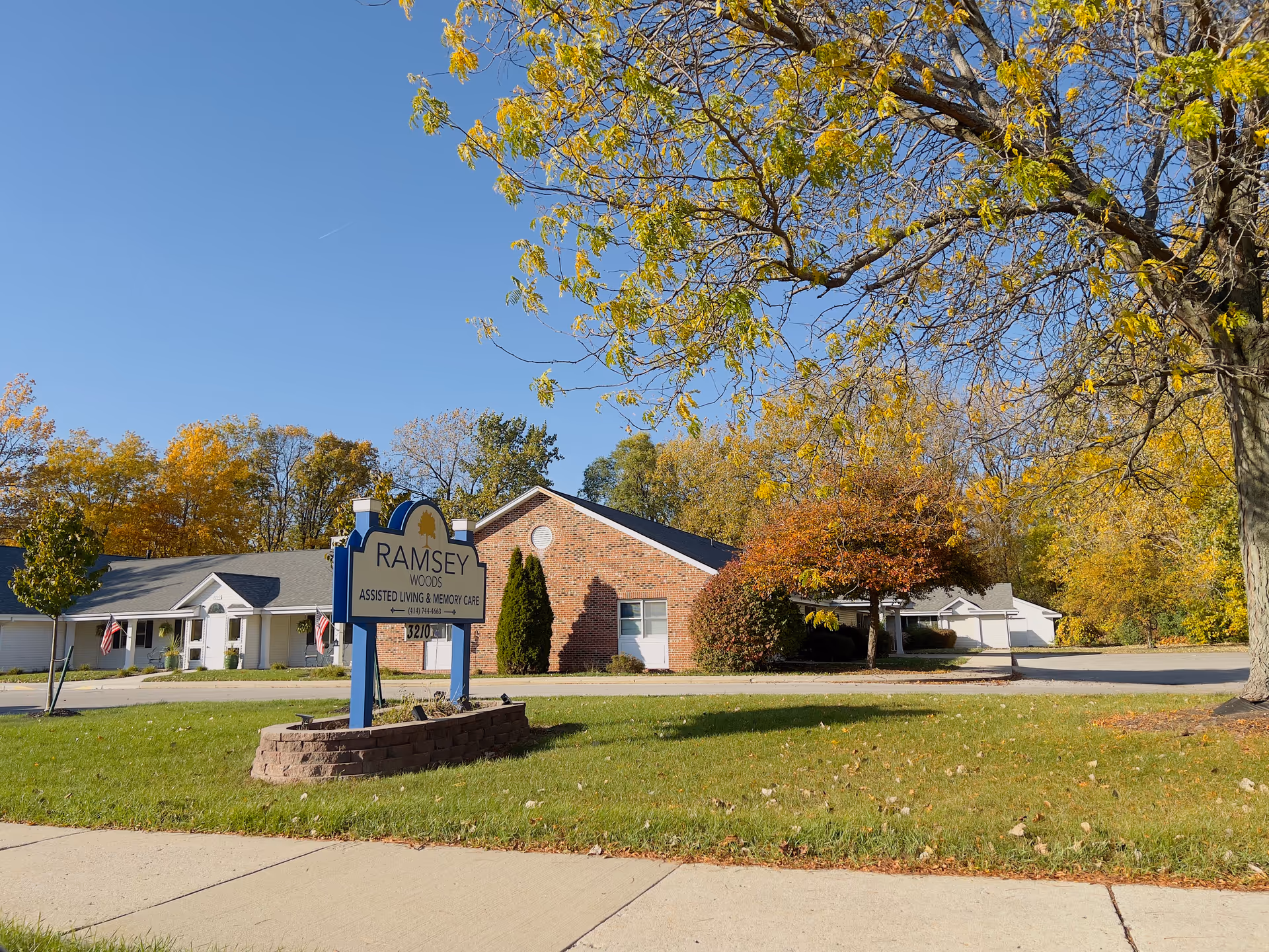 Exterior view of Ramsey Woods assisted living and memory care facility with a sign in the foreground on a grassy area. The building is single-story with brick and white siding, surrounded by trees with autumn foliage under a clear blue sky.