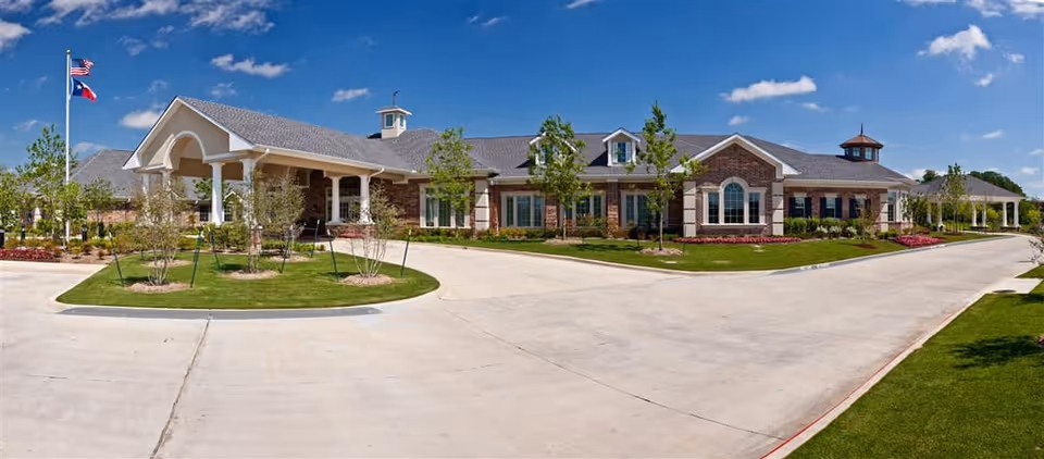 Front exterior of a single-story brick senior living facility with a circular driveway, landscaped lawns, and flagpoles under a blue sky.