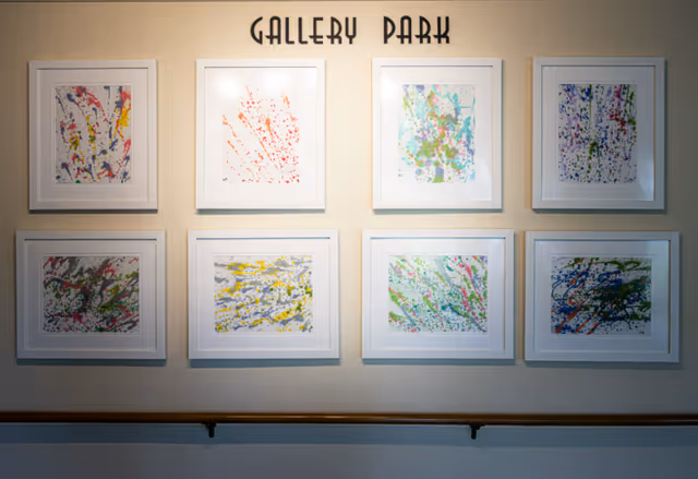 A wall display titled 'Gallery Park' featuring eight framed abstract paintings with colorful splatters and strokes arranged in two rows of four. Below the paintings is a wooden handrail attached to the wall.
