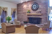 A cozy living room area with beige armchairs arranged around a stone fireplace. The fireplace has a wooden mantel decorated with a clock, flowers, and candles. The room features wooden flooring and windows with beige curtains and blinds.