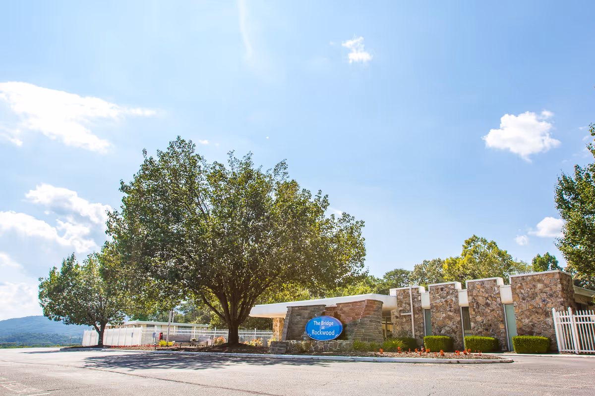Front exterior of Rockwood Care & Rehab Center with a low stone facade, large trees, a driveway, and a blue sign reading "The Bridge Rockwood".