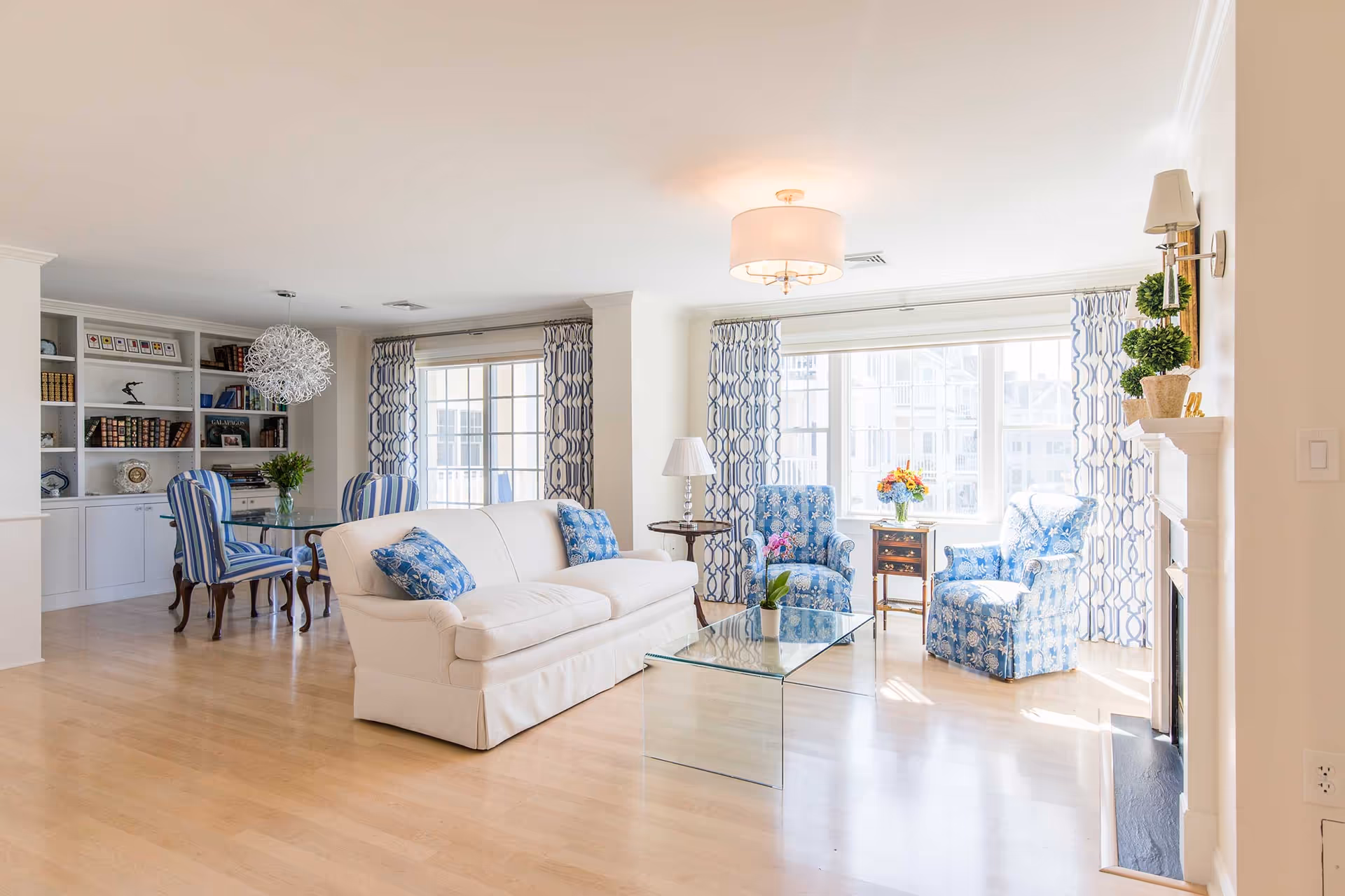 Bright and airy living room with light wood flooring, a white sofa with blue patterned pillows, two blue floral armchairs, a glass coffee table with a small plant, and large windows with blue and white patterned curtains. A dining area with a glass table and striped blue chairs is visible in the background, along with built-in white shelves filled with books and decorative items.