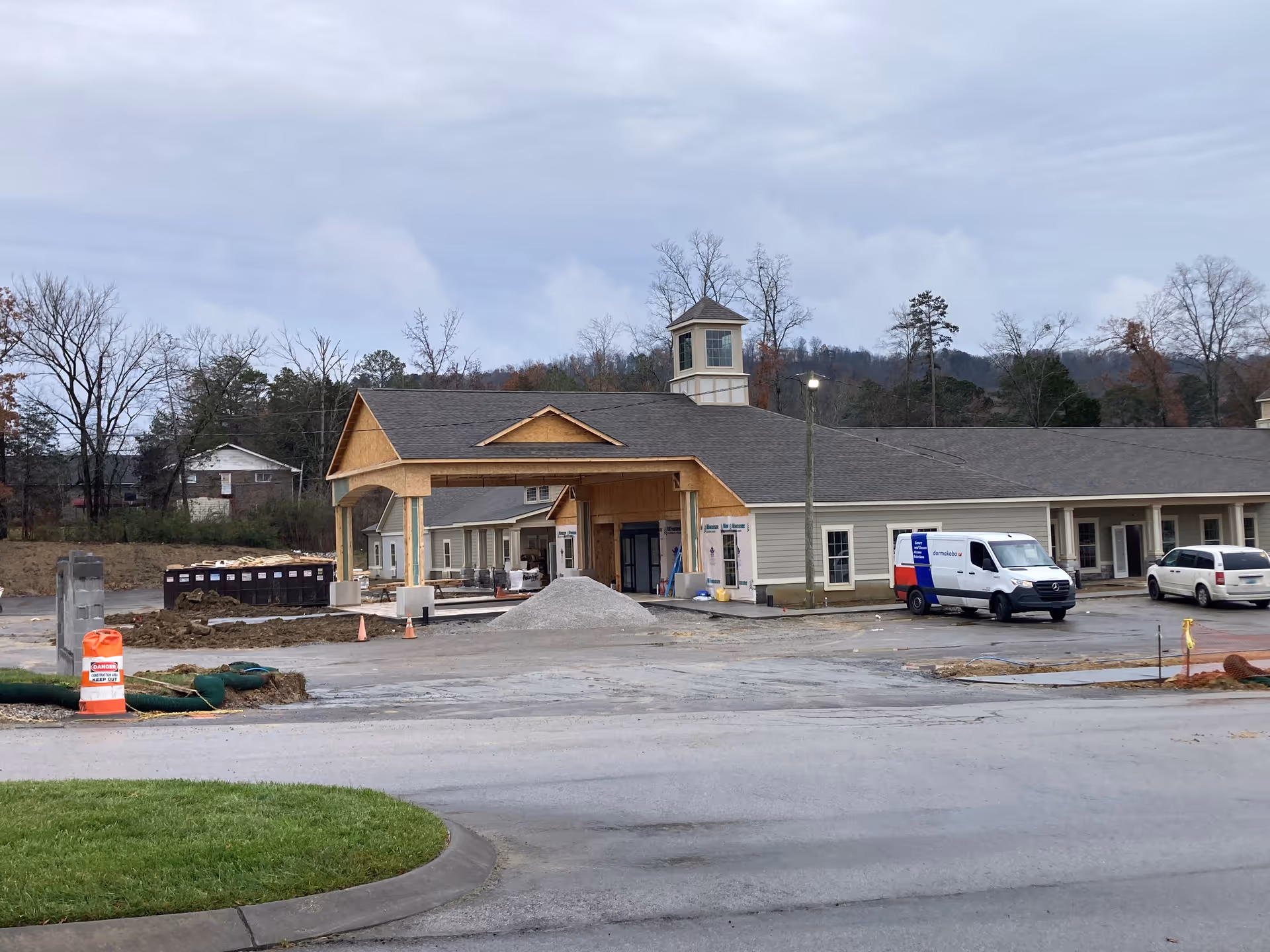 Exterior view of The Lantern at Morning Pointe Alzheimer's Center of Excellence, East Hamilton, showing a building under construction with a covered entrance, construction materials, and vehicles parked nearby. Trees and hills are visible in the background under a cloudy sky.