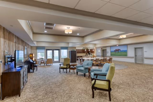 A spacious senior living common area with patterned carpet, multiple armchairs in green and blue, a wooden TV stand with a flat-screen TV, a piano, and a dining area with tables and chairs near a kitchen. Large windows and glass doors allow natural light to enter the room.