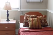 Single bed with patterned pillows and a red bedspread next to a wooden nightstand with a lamp in a small bedroom.
