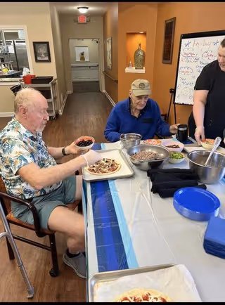 Two elderly men sitting at a table preparing personal pizzas with various toppings, while a staff member assists them. The setting appears to be a communal area in a retirement living facility with a whiteboard in the background.