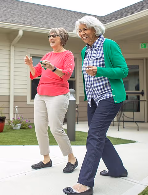 Two elderly women smiling and walking outdoors on a paved path in a courtyard area of a retirement community. One woman is wearing a pink top and light-colored pants, and the other is wearing a green cardigan over a blue and white checkered shirt with dark pants. The background shows the exterior of a building with windows and a door.
