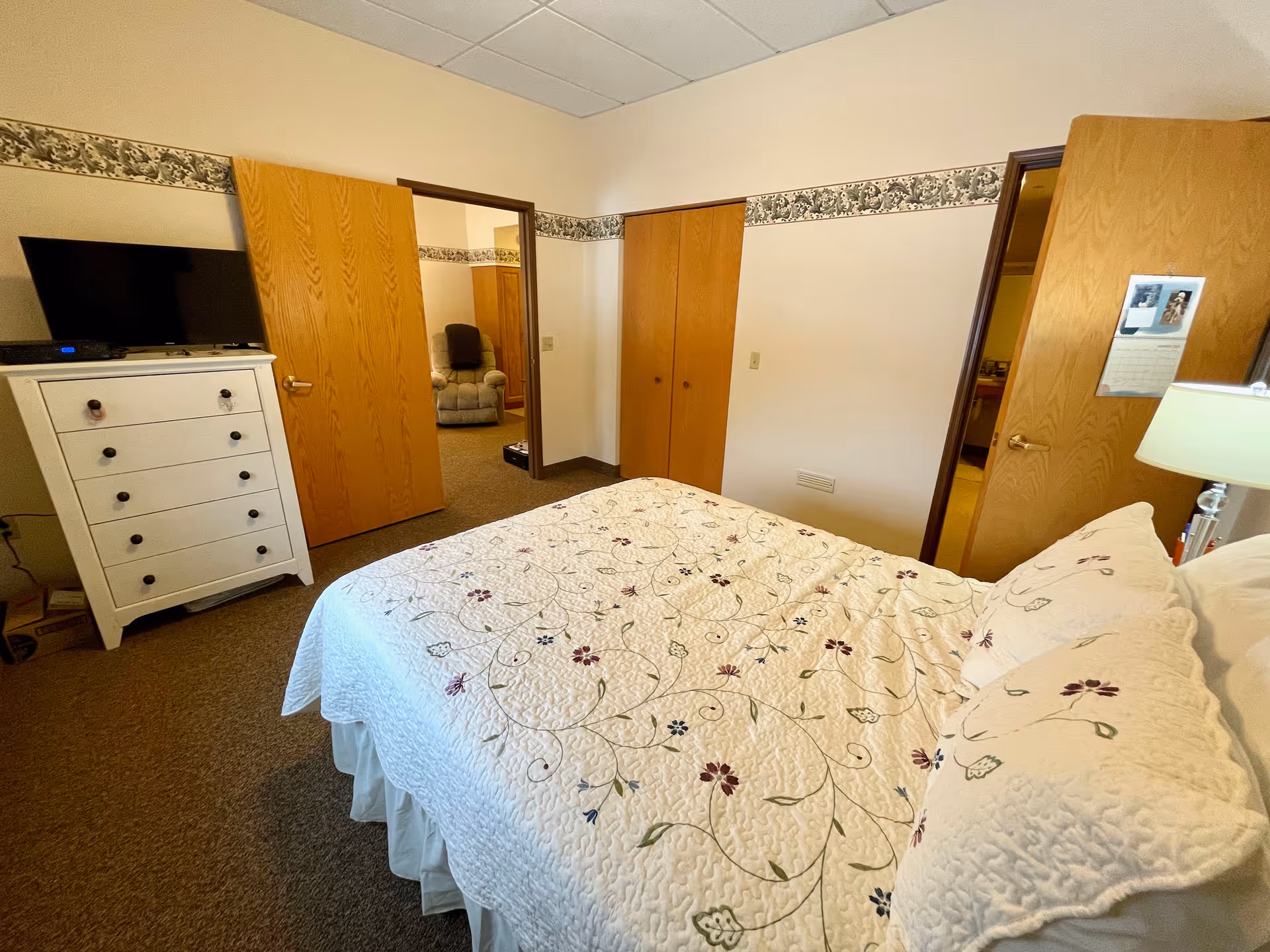 A bedroom with a bed covered in a white quilt with floral embroidery. There is a white dresser with a flat-screen TV on top against one wall. Two wooden doors are open, one leading to a room with a recliner chair and the other to a bathroom. The walls are white with a floral wallpaper border near the ceiling.
