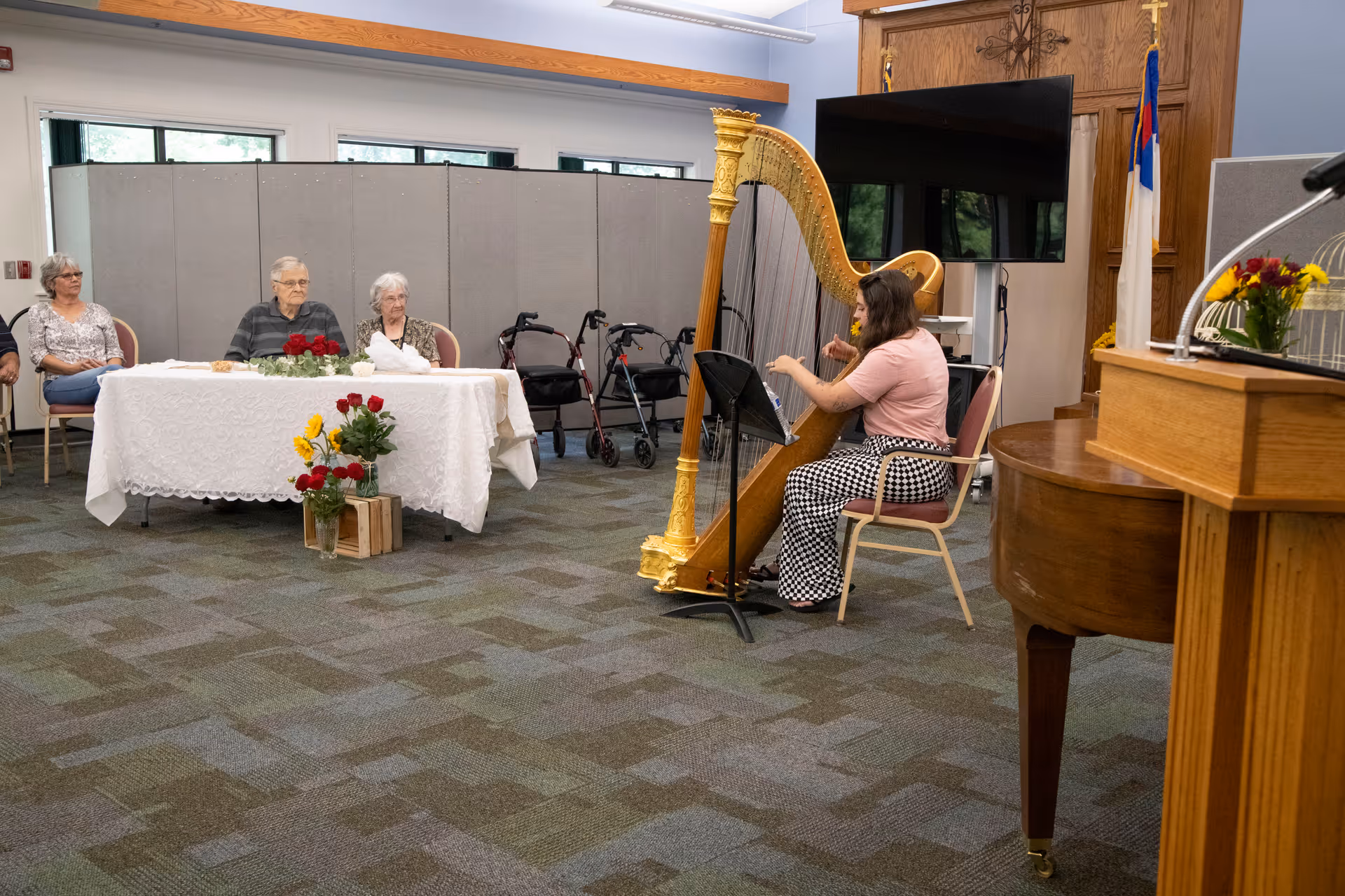 A woman is playing a large golden harp in a room with carpeted floor. Three elderly people are seated at a table covered with a white lace tablecloth, watching the performance. There are walkers behind the table and a large TV mounted on the wall. The room has wooden paneling and a podium with flowers on the right side.