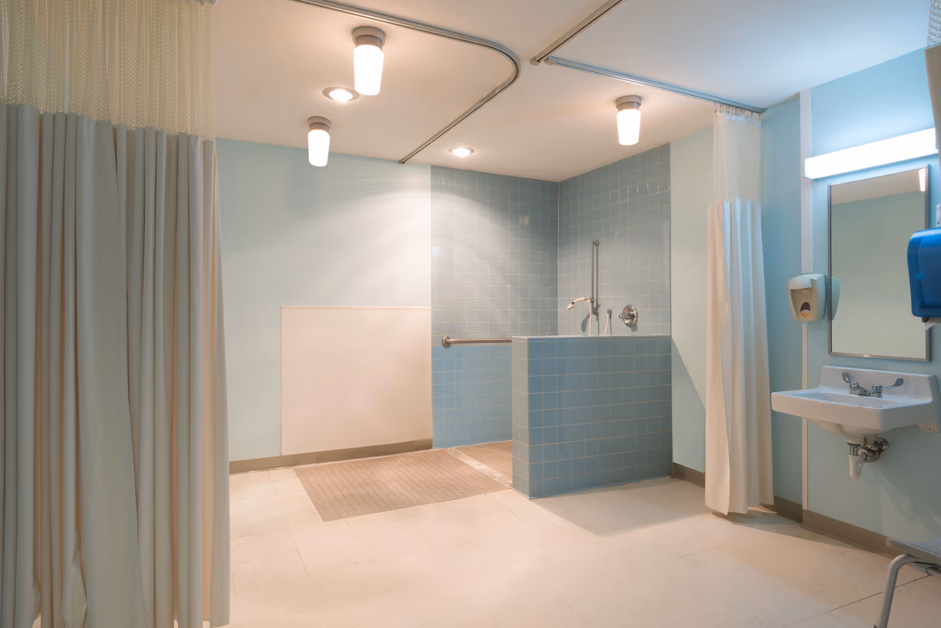 A clean, well-lit bathroom area with light blue tiled walls and a tiled shower section with a grab bar. There are ceiling lights and a curtain on a ceiling track for privacy. A white sink with a mirror and soap dispenser is mounted on the right wall.
