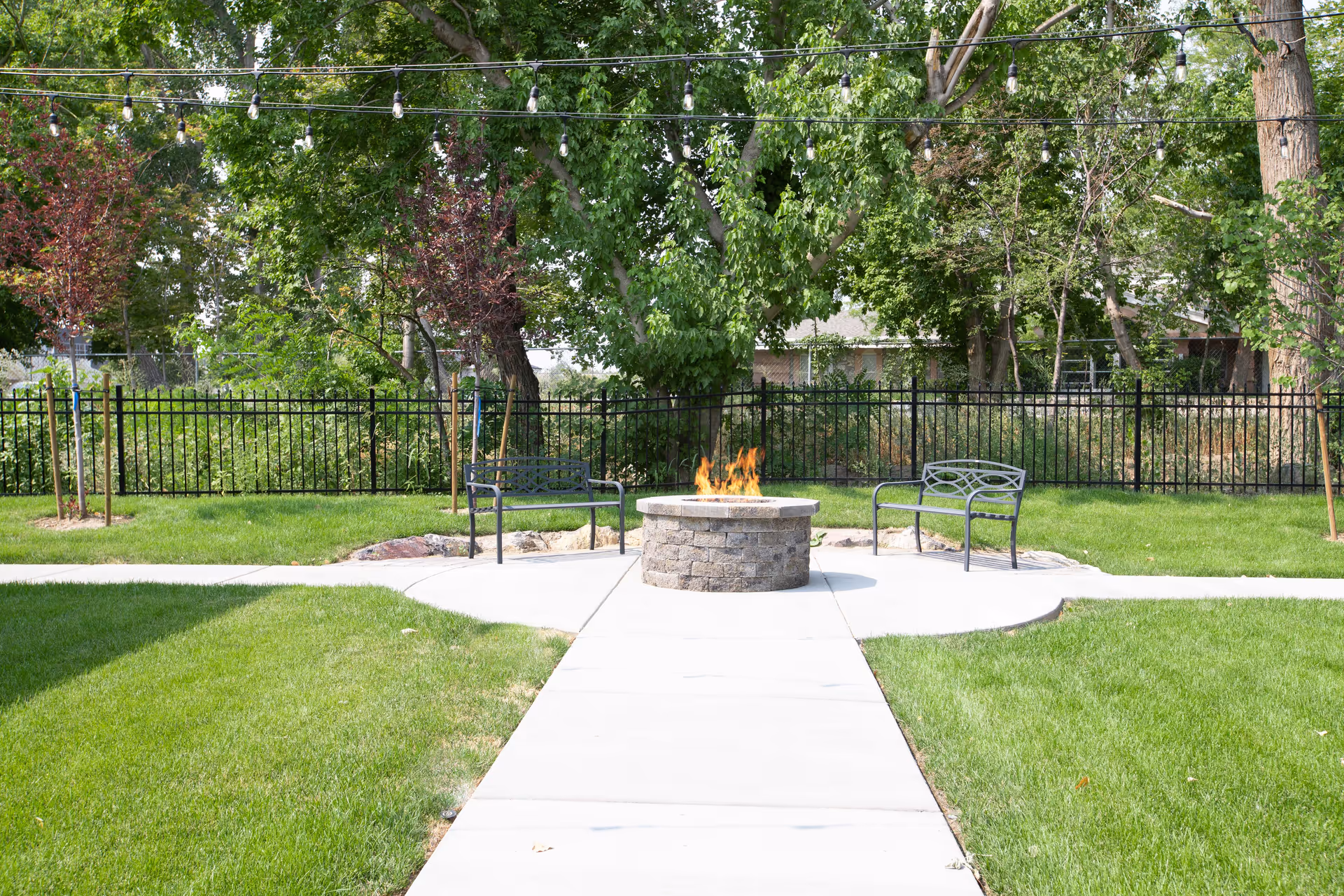 A grassy courtyard featuring a central stone fire pit with flames, two metal benches, string lights overhead, and trees and a black fence in the background.