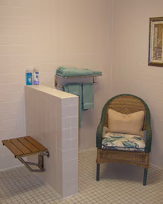 Tiled accessible shower area with a folding shower seat, towel rack with folded towels, and a wicker chair with cushions.