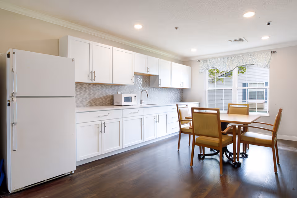 A bright kitchen area with white cabinets, a white refrigerator, a microwave on the countertop, and a wooden table with four chairs near a window with a floral valance.