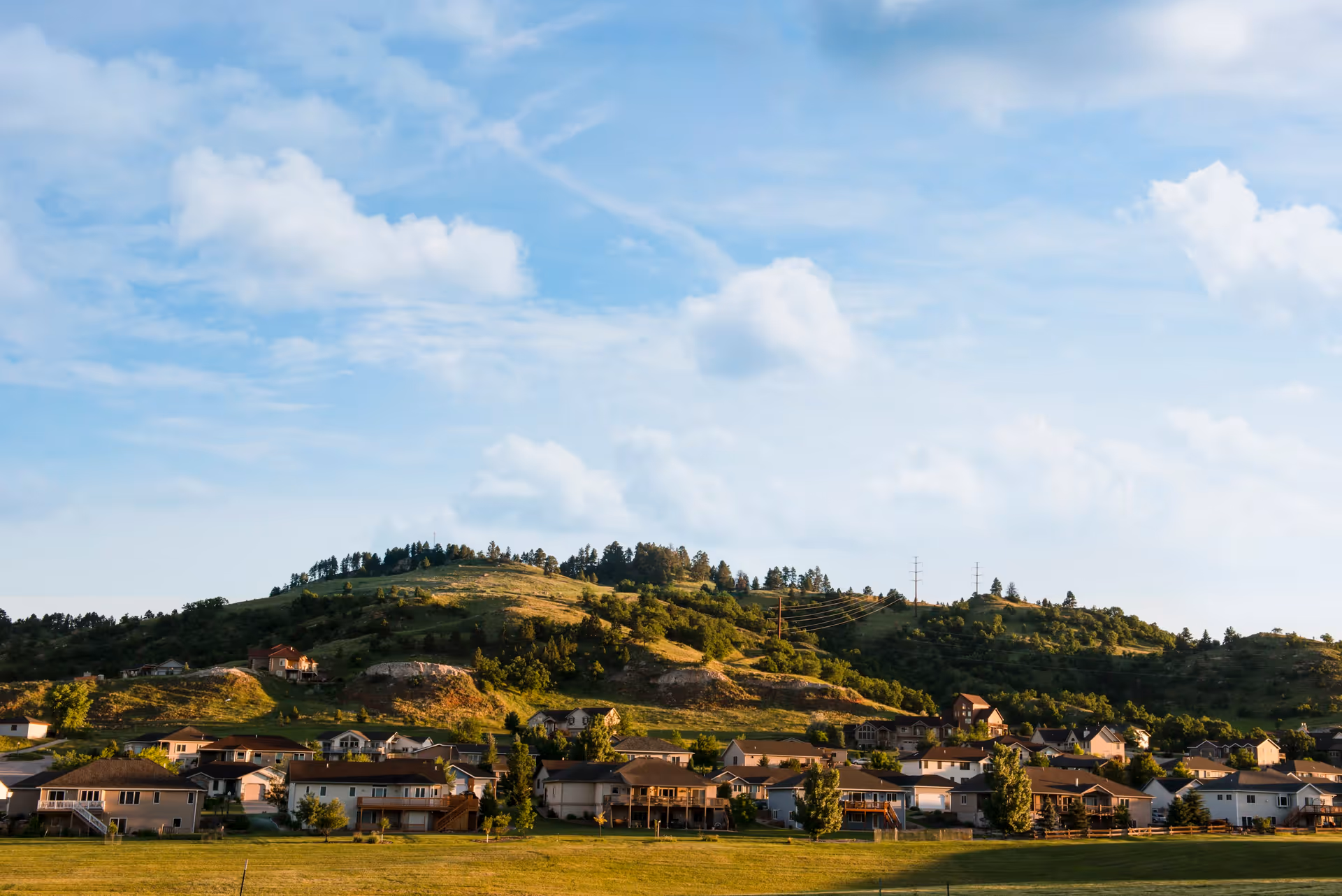 A scenic view of a residential neighborhood with houses at the base of a green, tree-covered hill under a partly cloudy blue sky.