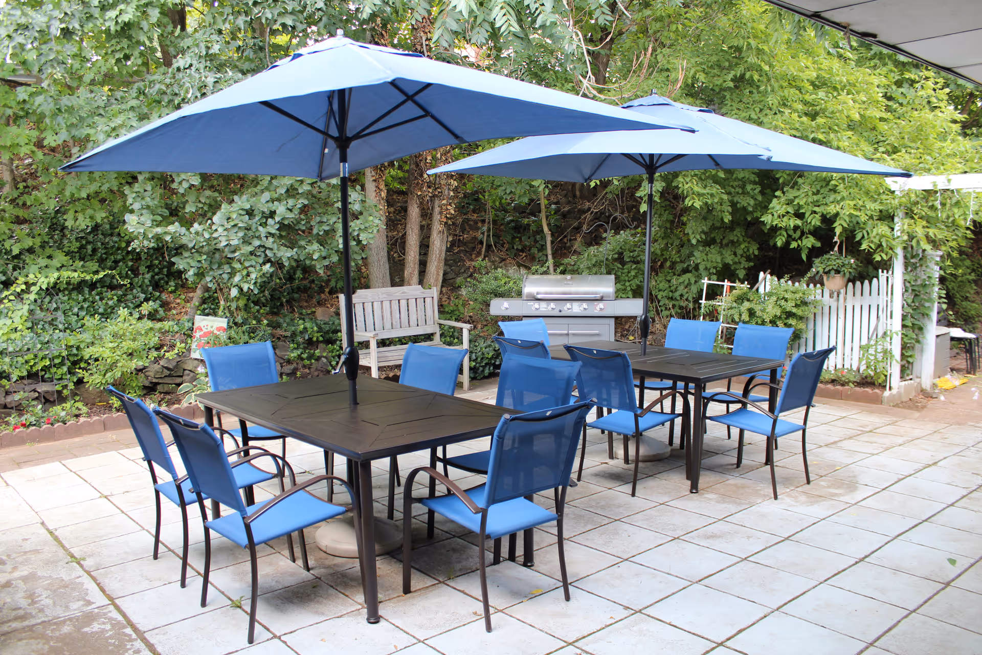 Outdoor patio with two dining tables, blue chairs and large blue umbrellas, a grill and bench surrounded by greenery.