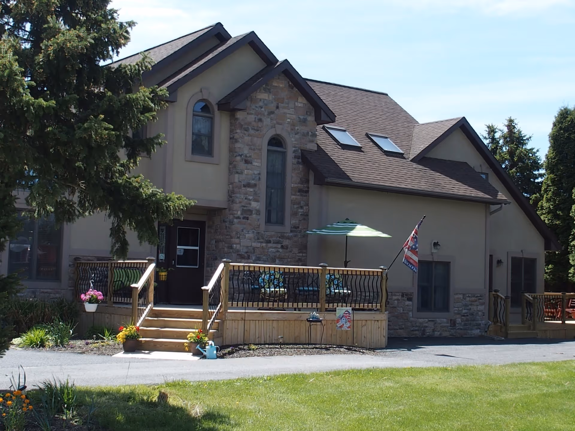 Exterior view of a two-story building with stone and beige siding, featuring a wooden deck with railings, outdoor seating, a green and white striped umbrella, and an American flag. The building is surrounded by trees, grass, and flower pots near the steps leading to the deck.