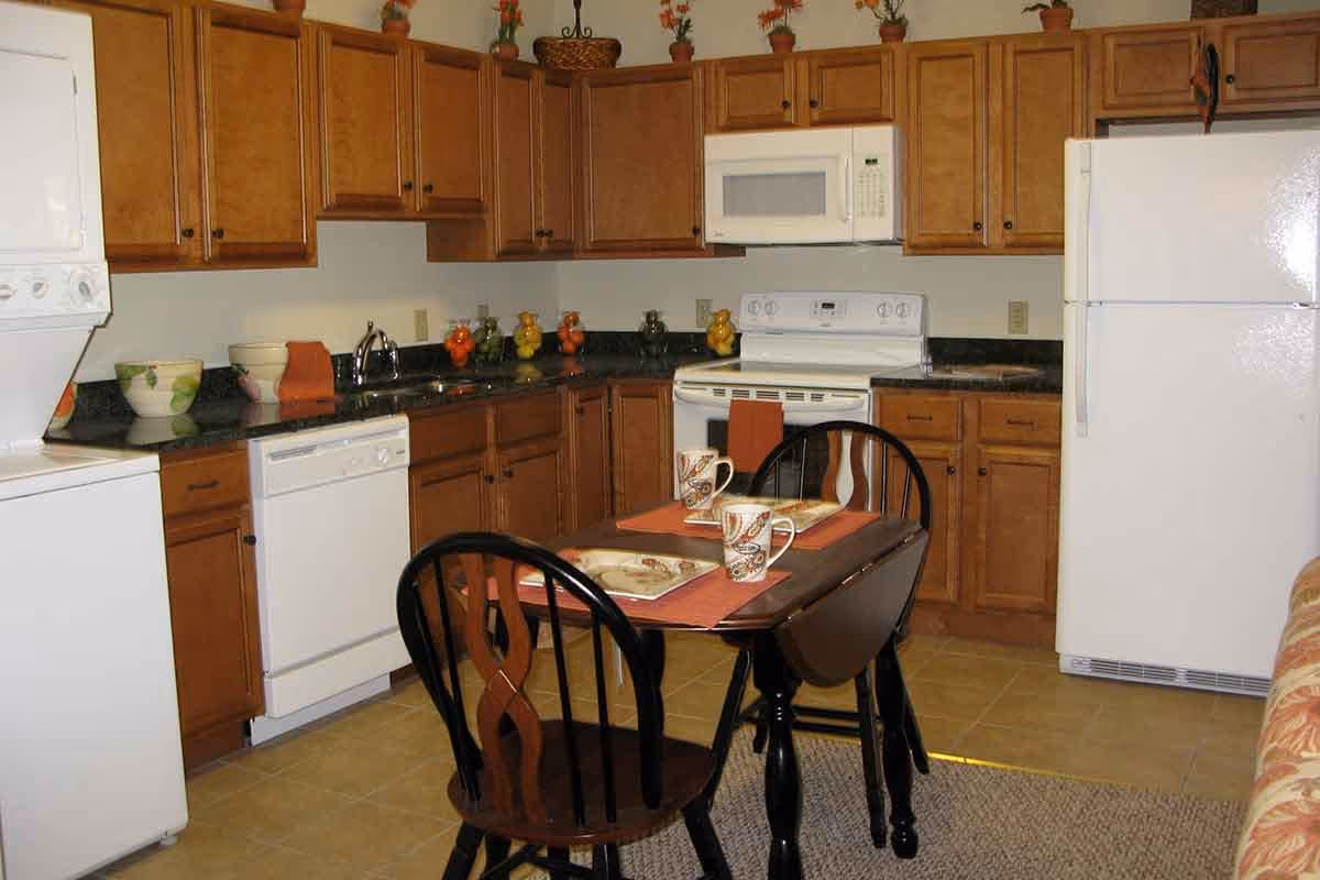 Compact kitchen with wooden cabinets, white appliances, and a small dining table set with two chairs.