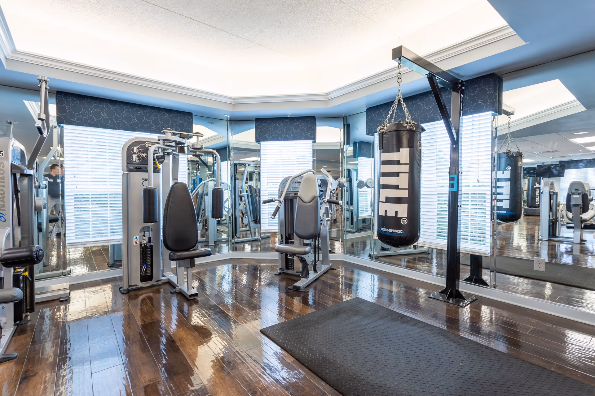 A bright fitness room with polished wooden floors, mirrored walls, and large windows with blinds. The room contains exercise machines and a hanging TITLE punching bag on a black stand. A black exercise mat is placed on the floor.