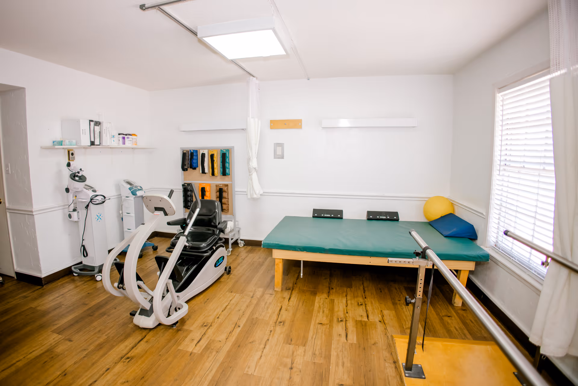 A physical therapy room with exercise equipment including a recumbent exercise bike, a padded therapy table with a yellow exercise ball and blue cushion, parallel bars for walking support, and various therapy supplies on shelves and racks. The room has wooden flooring and white walls with a window covered by blinds.