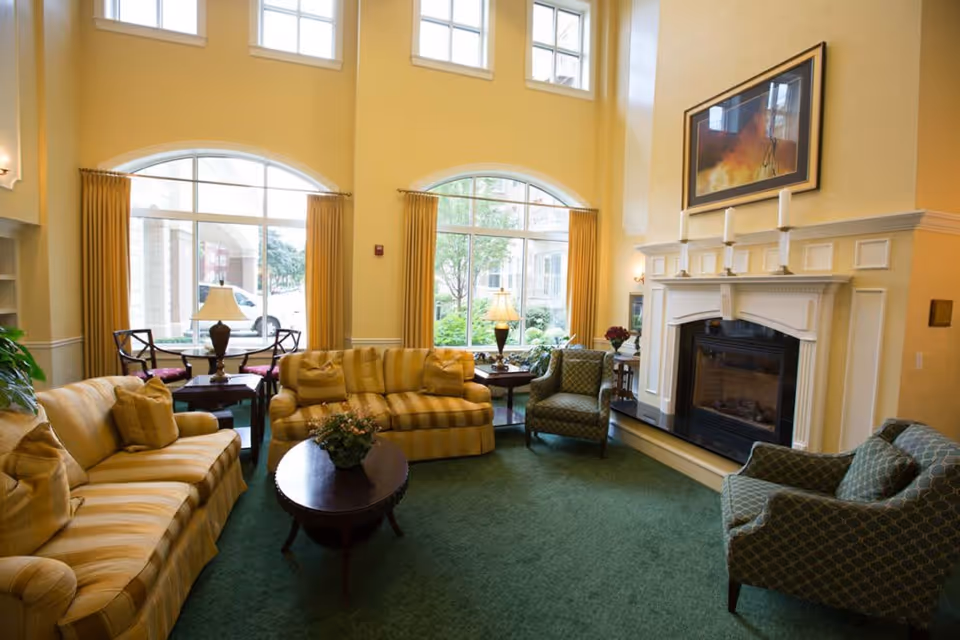 A cozy living room area with yellow striped sofas, two patterned armchairs, a wooden coffee table with a plant, and a fireplace with candles on the mantel. Large windows with yellow curtains let in natural light, and there are table lamps and chairs near the windows.