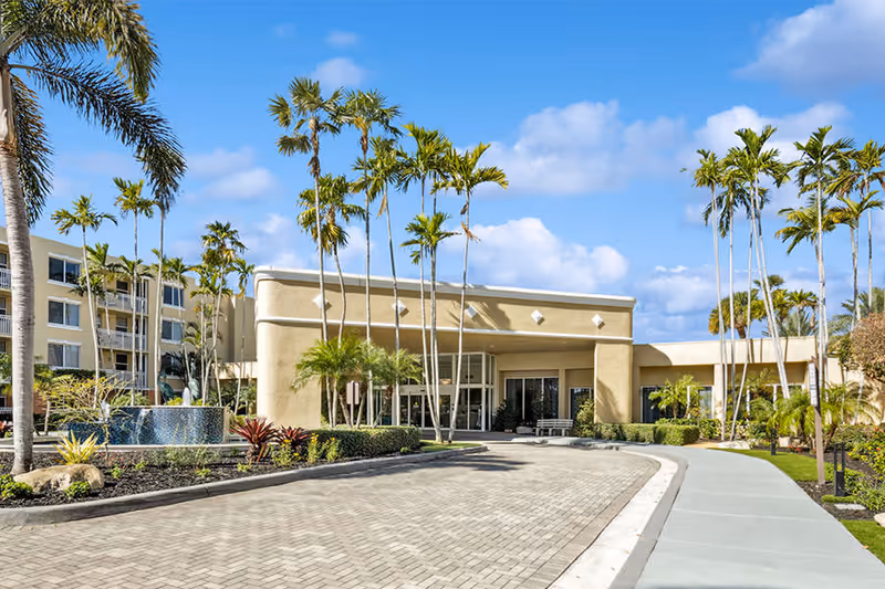 Exterior view of a senior living facility building with a driveway, palm trees, and landscaped greenery under a blue sky with some clouds.