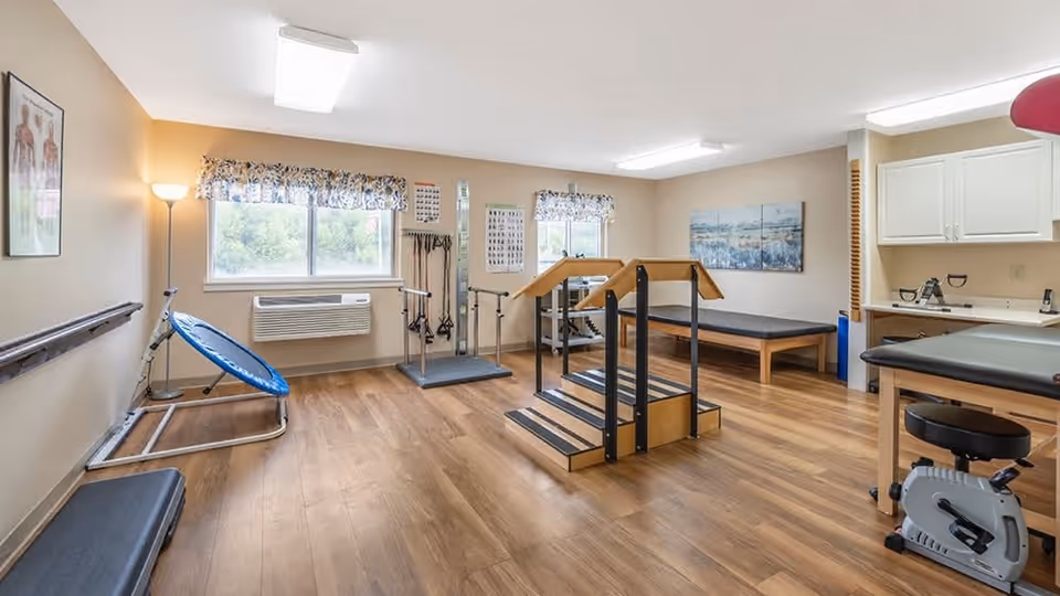 A physical therapy room with wooden flooring, exercise equipment including a small trampoline, parallel bars with steps, a stationary bike, and therapy tables. The room has two windows with floral valances, beige walls, and medical posters on the walls.