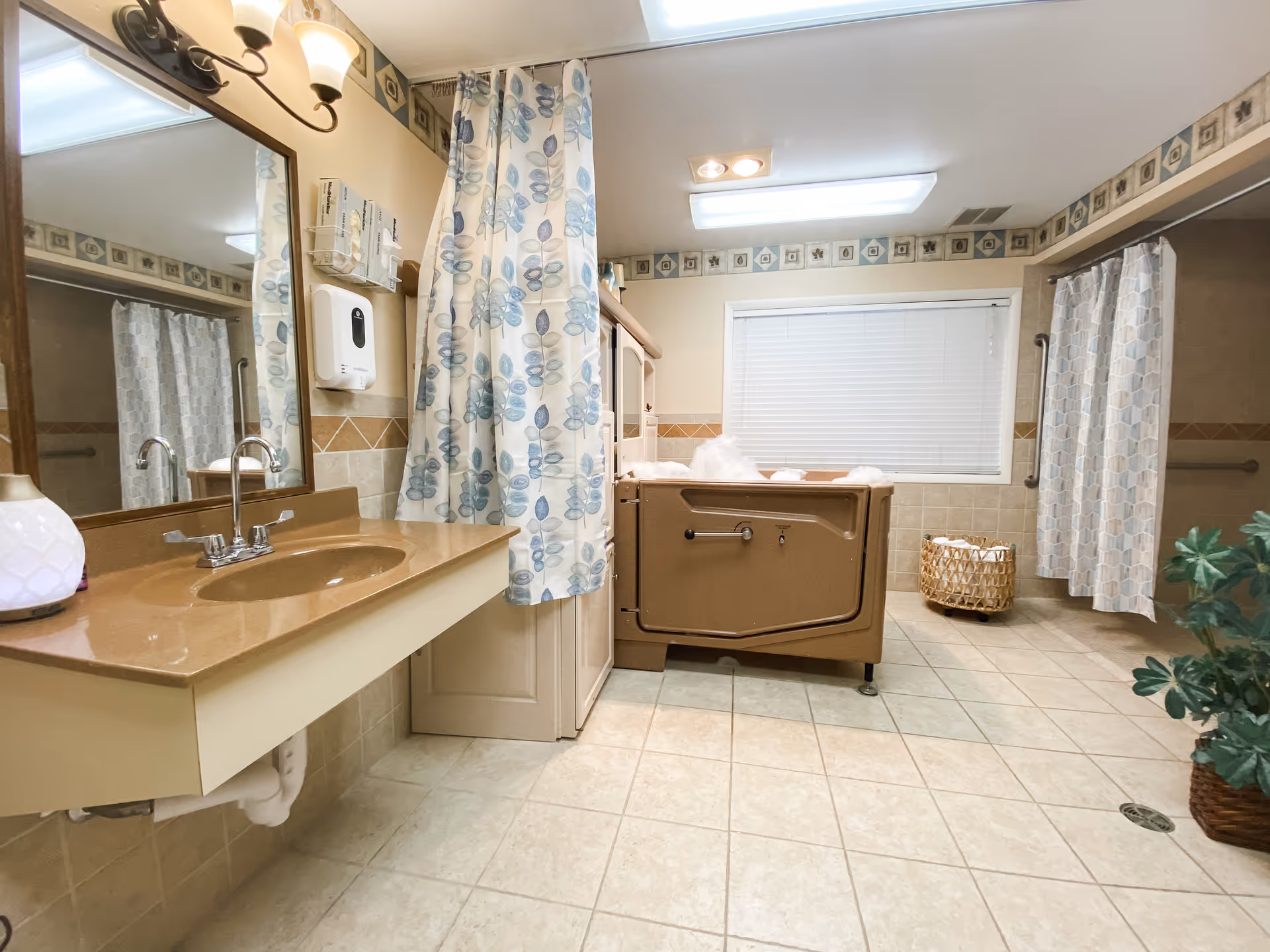 A spacious bathroom with beige tiled floor and walls, featuring a brown walk-in bathtub filled with bubbles, a beige countertop with a sink and faucet, a large mirror above the sink, floral patterned shower curtains, a wicker basket with towels, and a potted plant in the corner.