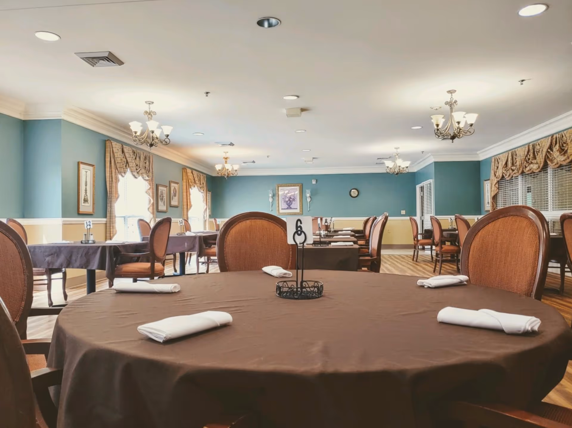 Spacious dining room with round tables draped in brown tablecloths, rolled napkins, upholstered chairs, chandeliers, and teal walls with windows.