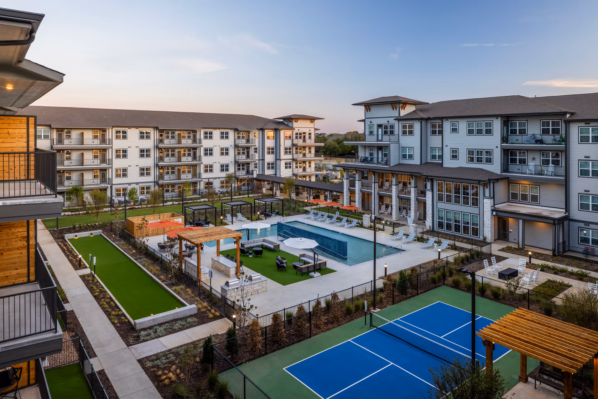 View of an outdoor recreational area in a senior living facility named Solea Lewisville, featuring a swimming pool surrounded by lounge chairs and umbrellas, a bocce ball court, a blue tennis or pickleball court, pergolas with seating, and multi-story residential buildings in the background under a clear sky at dusk.