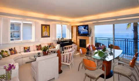 Bright living and dining area with sofas, a piano, fireplace and a glass dining table, opening to a balcony with ocean views.