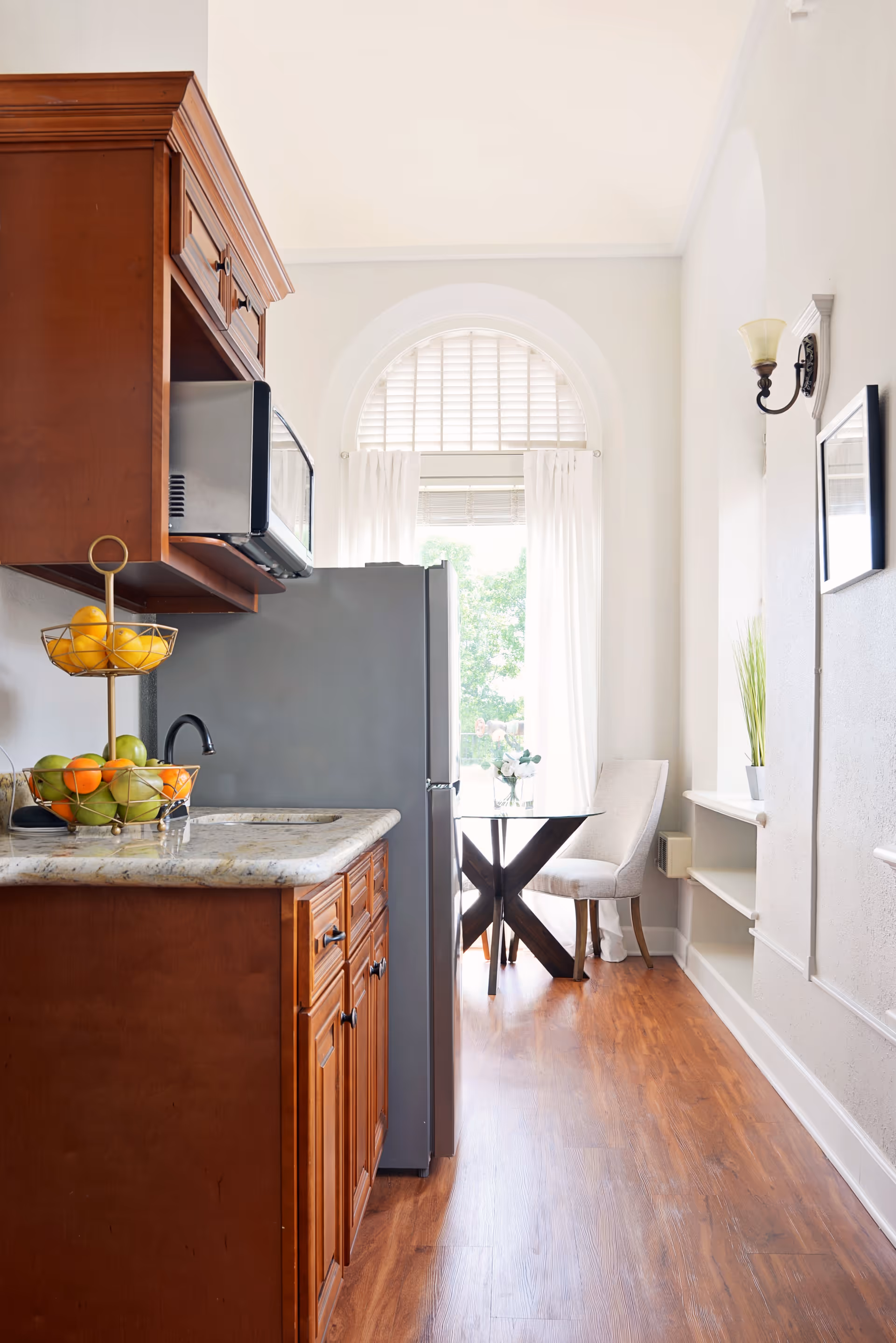 Bright narrow kitchen with wooden cabinets and a marble countertop leading to a small dining table by an arched window with white curtains.